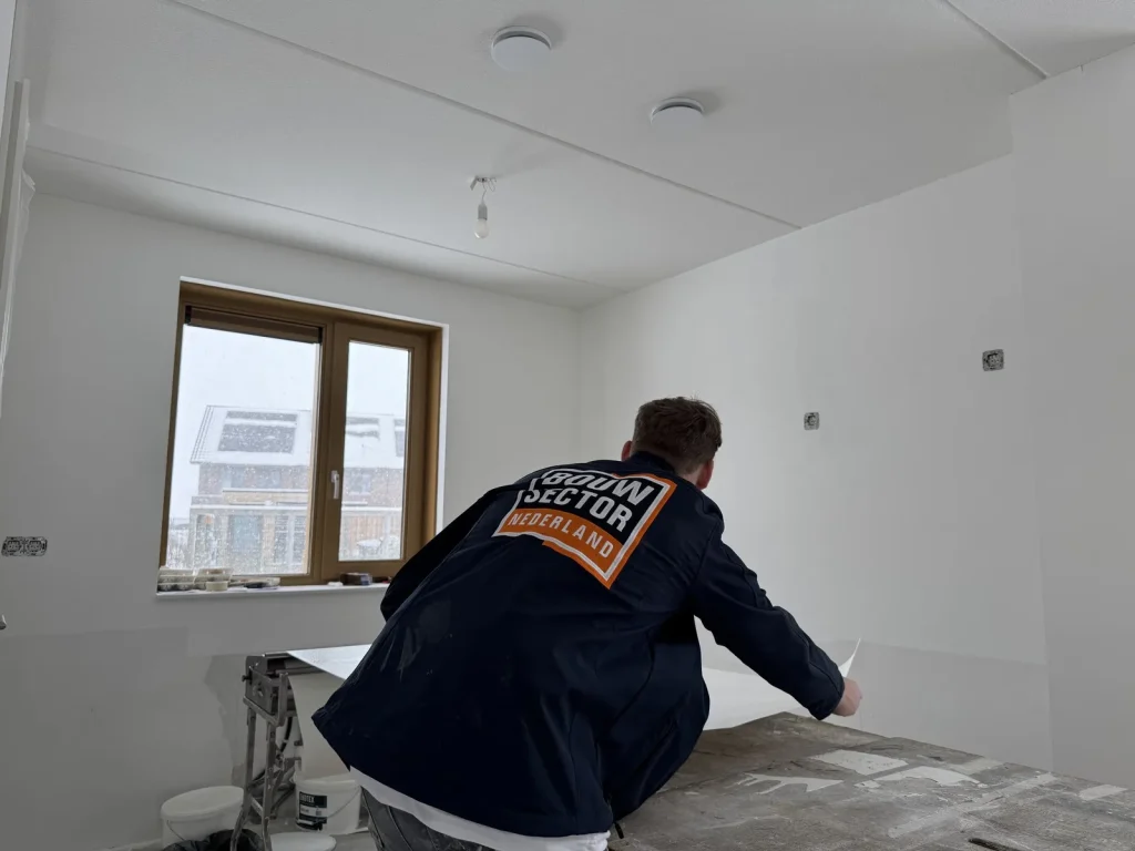 Behanger van Bouwsector Nederland rolt een baan vlies uit over de plaktafel in een kamer met houten kozijnen